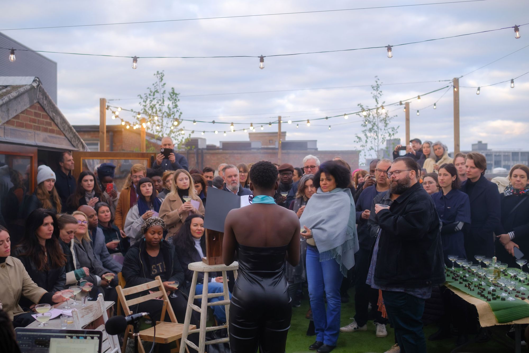 Image of the roofterrace with audience gathered around performer during Zina Saro Wiwa's Illicit Gin Assembly #4